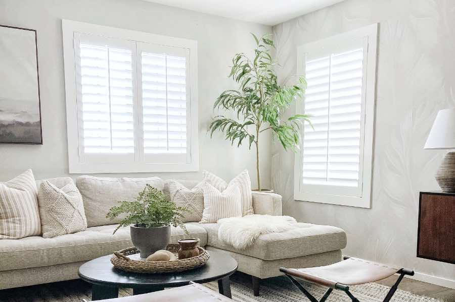 Neutral-colored window treatments help your windows blend in with your decor so their size difference doesn't stand out. White Polywood shutters on windows of different sizes in a living room